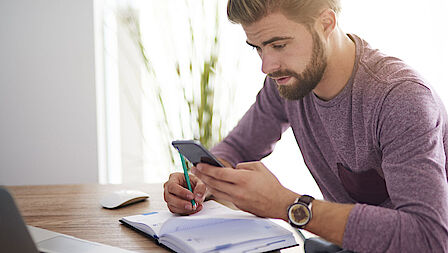 Een man zit met een schrift en ene laptop aan tafel met een rekenmachine in zijn hand.