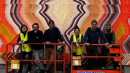 Een groep mensen staat voor een kleurrijke muurschildering. Onder hen de twee kunstenaars die in het artikel worden genoemd.