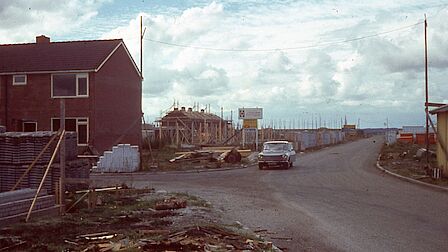  Een historische foto van de wijk Barnewiel in Oudkarspel tijdens de bouw in de jaren ‘70. Op de voorgrond is een deels afgebouwde woning te zien, omringd door bouwmaterialen. Een klassieke auto rijdt over een onverharde weg, terwijl op de achtergrond meerdere huizen in aanbouw zijn met steigers en houten constructies.