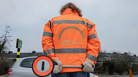 Een klaar-over met een oranje jas aan houdt een stopbordje op de rug vast.