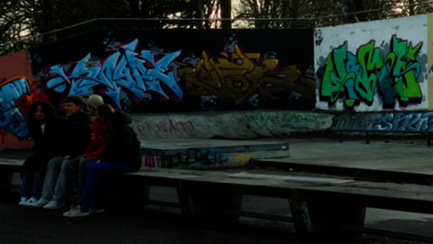 Op deze header zie je een lange betonnen bank met daarop drie jongeren. Achter hen zijn borden met grafitti zichtbaar met diverse teksten in licht- en donkerblauw en groen. Lastig leesbaar ...