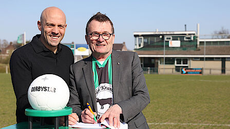 De voorzitter en de wethouder ondertekenen lachend de overeenkomst. OP het tafeltje voor hen ligt een wedstrijdbal.