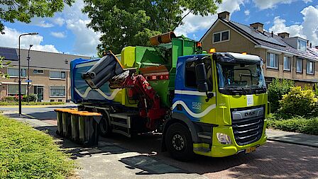 Vuilniswagen van gemeente Dijk en Waard leegt PMD-afvalbakken met oranje deksel in woonwijk. 