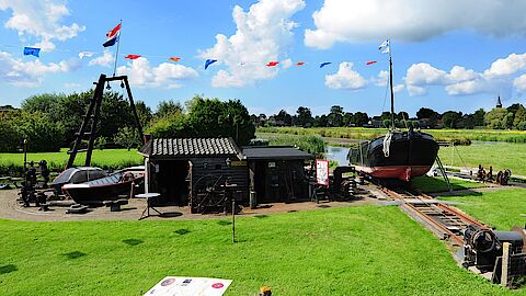 Overzicht van een museale schuitenhelling, met gebouw, schuit en zonnige lucht met wolken.